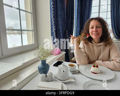 Donna seduta a un tavolo con un dessert davanti a sé. Sorride e mangia un pezzo di torta al ristorante Foto Stock