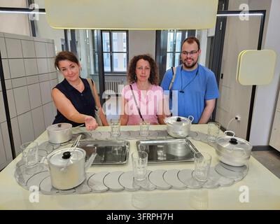 Tre persone sono in piedi davanti a un tavolo con uno sfondo giallo da cucina. Sul tavolo ci sono diverse tazze e ciotole con il cartello Arte. Foto Stock