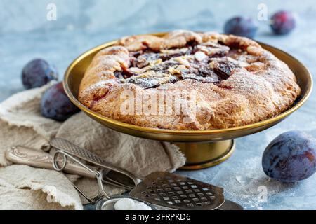 Supporto per torte in bronzo con ripieno di prugne e scaglie di mandorle e prugne blu fresche su un tavolo di cemento strutturato Foto Stock