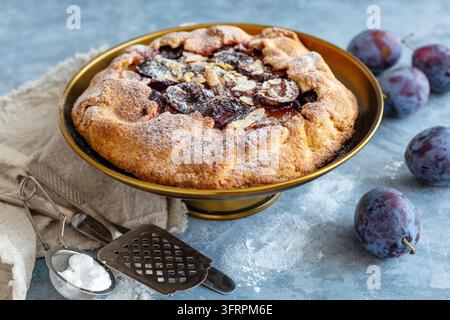 Supporto per torte in bronzo con ripieno di prugne e scaglie di mandorle e prugne blu fresche su un tavolo di cemento strutturato Foto Stock