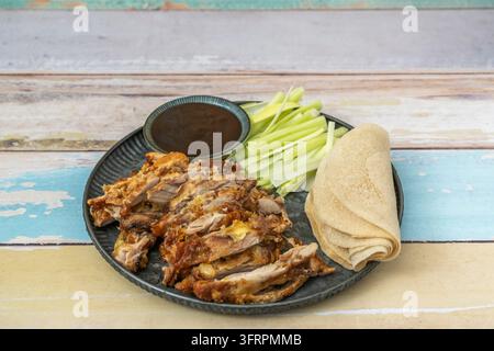 Fette di anatra croccanti servite con pancake sottili, bastoncini di scalogno e salsa da intingere su un piatto scuro sopra legno colorato. Foto Stock