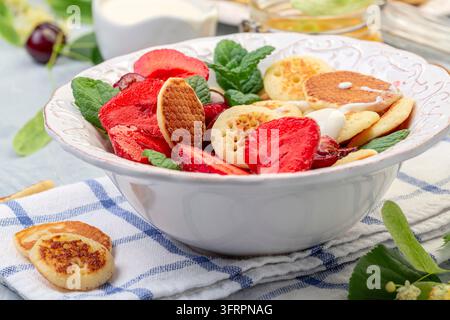 Piccoli pancake con salsa di miele e panna, fragole e ciliegie serviti con menta fresca in una ciotola di ceramica bianca, messa a fuoco selettiva. Mini pancake alla moda Foto Stock