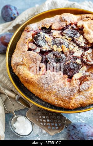 Supporto per torte in bronzo con ripieno di prugne e scaglie di mandorle e prugne blu fresche su un tavolo di cemento strutturato Foto Stock
