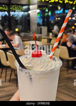 Persona che tiene una tazza di gelato o milkshake al cocktail con una ciliegia in cima. La ciliegia è su una cannuccia in un caffè Foto Stock