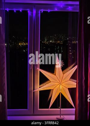 Una grande decorazione a forma di stella illuminata è posizionata su un supporto davanti a una finestra. La finestra è leggermente aperta, permettendo alla luce della decorazione di filtrare e illuminare la stanza Foto Stock