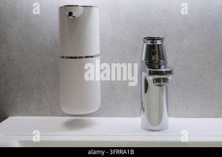 Primo piano di un erogatore di sapone automatico bianco montato su una parete grigia vicino a un rubinetto dell'acqua cromato lucido sopra un lavandino in ceramica. Moderno minimal Foto Stock