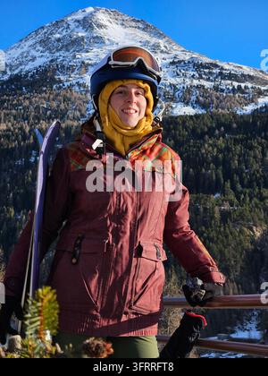 Una donna con un cappotto rosso e occhiali protettivi è in piedi su una ringhiera con uno snowboard. Sorride e sta godendo la vista della montagna nella stazione sciistica Foto Stock