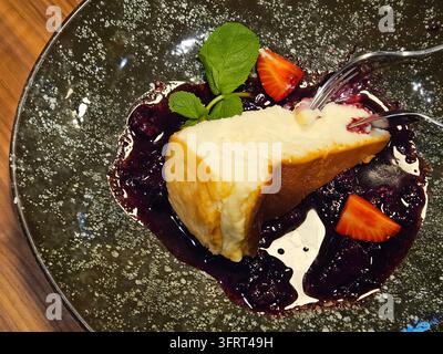 Fetta di cheesecake con fragole e menta sopra. Il piatto è nero e il dessert è su una forchetta Foto Stock