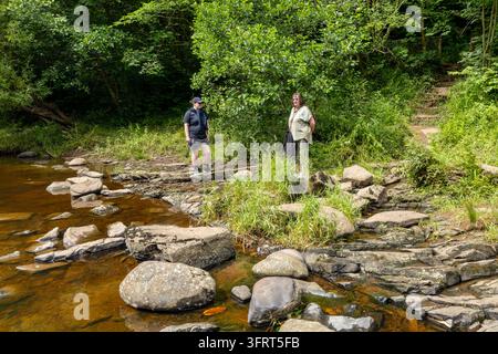 Due donne di generazioni diverse stanno su rocce di arenaria piatte al margine poco profondo del fiume, circondate da rigogliosi alberi decidui verdi e rive selvatiche Foto Stock