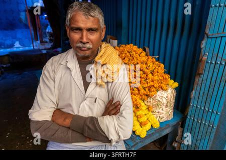 Un uomo dai capelli grigi con i baffi e un panno giallo sulla spalla si trova a braccia incrociate accanto a calendule arancioni ammucchiate al mercato dei fiori, Noi Foto Stock