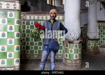 Un uomo con un gilet di maglia e jeans con una borsa a tracolla tiene un mazzo di rose rosse mentre è in piedi tra muri piastrellati verdi e colonne bianche Foto Stock