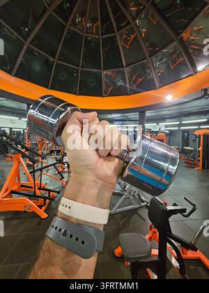 Mano afferra un manubrio in un contesto luminoso di palestra. Attrezzature arancioni e soffitto in vetro incorniciano l’inquadratura. Il manubrio riflette vivacemente le luci della palestra. La persona indossa un fitness tracker al polso Foto Stock