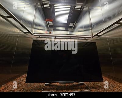 Mobiletti TV moderni in un interno di ascensore metallico. Le superfici riflettenti mostrano una sottile illuminazione ambientale. Lo schermo scuro contrasta con il pavimento ruvido. Linee pulite e design industriale dominano la vista Foto Stock