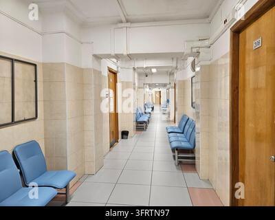 Mosca – 24 ott. 2025: Lungo corridoio dell’ospedale con sedili blu e pareti piastrellate. Soffitto chiaro e porte di legno allineano il corridoio. Nessun animale o pianta è visibile in questa immagine Foto Stock