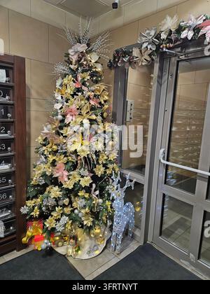 Albero di Natale decorato con luci e ornamenti. L’albero è accanto alle porte di vetro in un atrio. Una ghirlanda festosa adorna la porta sopra. Decorazioni dorate e rosa illuminano l’albero Foto Stock