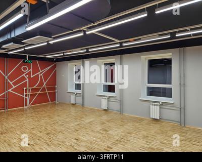 Mosca - 6 dic. 2025: Studio di danza vuoto con pavimento in legno e luci moderne al soffitto. Finestre bianche costeggiano le pareti grigie con radiatori sottostanti. Parete rossa con design geometrico bianco astratto. Spazio Foto Stock