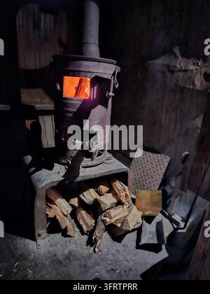 Stufa a legna rustica risplende di calore. I tronchi sono impilati ordinatamente accanto. Pareti di legno scuro incorniciano la scena. Atmosfera accogliente di capanna debolmente illuminata. La luce del fuoco proietta un bagliore soffuso e tremolante Foto Stock