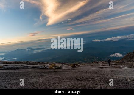 Escursionista solitario si trova in cima a una cima montuosa frastagliata. Nuvole lontane si muovono su un vasto orizzonte. La luce del sole bagna il cielo in morbide tonalità dorate. Il terreno è roccioso Foto Stock