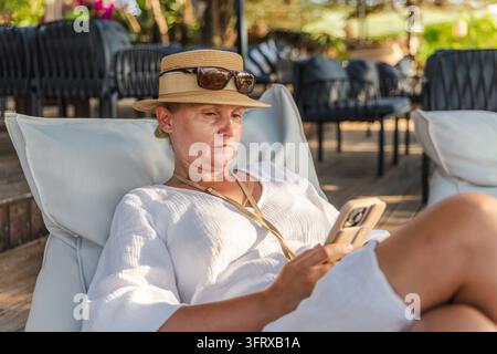 Donna con cappello da sole si rilassa su una sdraio all'aperto mentre utilizza uno smartphone. Foto Stock