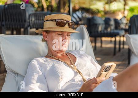 Donna con cappello da sole si rilassa su una sedia da esterno mentre utilizza il suo smartphone. Foto Stock
