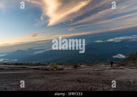 Escursionista solitario si trova in cima a una cima montuosa frastagliata. Nuvole lontane si muovono su un vasto orizzonte. La luce del sole bagna il cielo in morbide tonalità dorate. Il terreno è roccioso Foto Stock