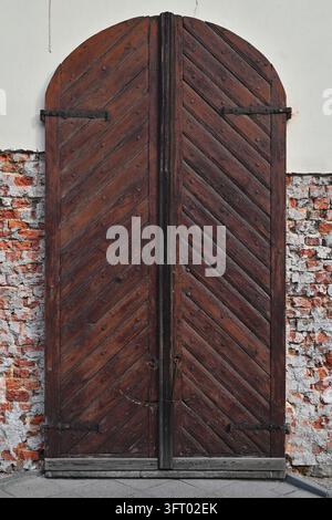 Ampia porta doppia in legno ad arco con motivo a spina di pesce inserita in un muro di mattoni ruvidi con residui di intonaco. Foto Stock