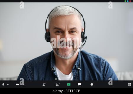 Uomo anziano sorridente che ha una videoconferenza mentre si è a casa durante la pandemia COVID-19, utilizzando cuffie, ritratto di primo piano, schermo pov. Tecnologia moderna Foto Stock