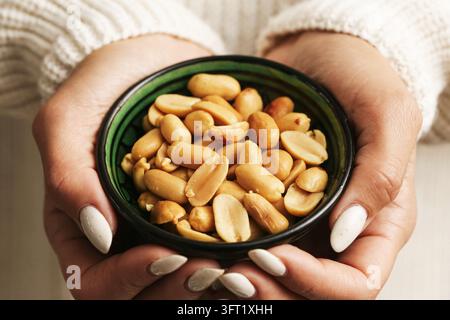 Donna che tiene una ciotola verde di arachidi sgusciate con entrambe le mani. Sfondo di snack sano per noci salate, cibi ricchi di proteine e mangiare informale. Foto Stock