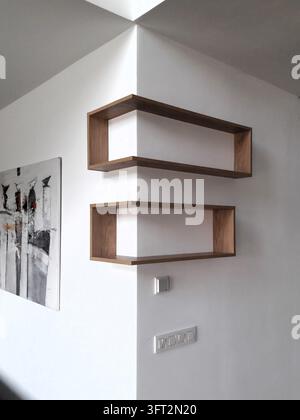 Una fotografia architettonica d'interni che mostra scaffali angolari in legno moderni e unici progettati per adattarsi intorno all'angolo sporgente di una parete bianca in un con Foto Stock