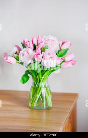 Tulipi rosa e peonie disposte elegantemente in un vaso trasparente su un tavolo di legno in un ambiente luminoso. Foto Stock