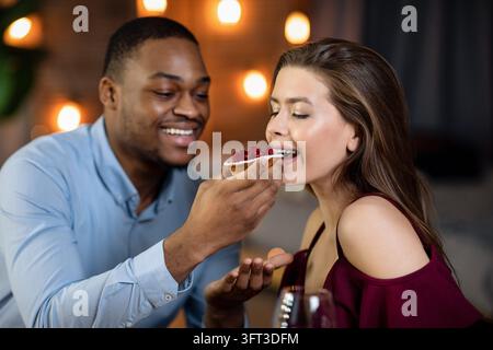 Allegra coppia interrazziale che si diverte a condividere il dessert insieme durante il romantico appuntamento al ristorante, amare l'uomo afroamericano che dà da mangiare al suo felice Girfri Foto Stock
