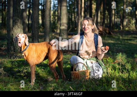 Donna allegra cacciatrice di funghi che passa il tempo libero con il cane all'aperto nella foresta raccogliendo porcini, escursionista felice che posa con l'animale domestico e mostra un fungo boletus, stile di vita attivo e vita naturale Foto Stock