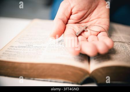 Un palmo aperto mostra un piccolo ciondolo a forma di croce d'argento e la sua catena sopra le pagine di un libro aperto, creando un primo piano orizzontale che enfatizza un Chr Foto Stock