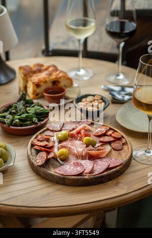 Prima vista di un vassoio di legno rustico con carni affumicate, chorizo, prosciutto e olive verdi servite con vino bianco. Foto Stock
