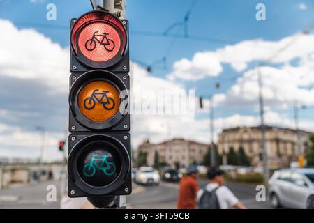Il semaforo per il traffico ciclabile brilla di rosso, ambra e verde su una strada di Budapest con traffico sfocato, pedoni ed edifici storici. Foto Stock