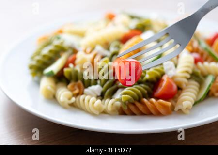 Primo piano di un'insalata di pasta fusilli tricolore colorata con pomodorini, zucchine e formaggio bianco su un piatto. Forchetta che tiene un pomodorino nella fo Foto Stock