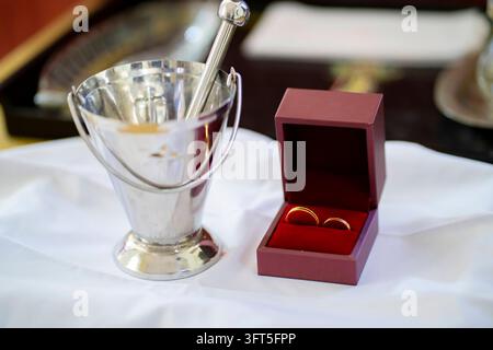 Anelli nuziali d'oro in una scatola accanto ad un aspergillo per l'acqua santa per la cerimonia religiosa Foto Stock