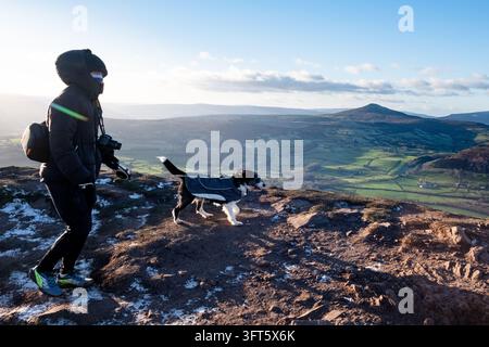 PERSONA CHE PASSEGGIA UN CANE SU UN SENTIERO MOUNTAIN, SENTIERO DI ESCURSIONISMO SKIRRID MOUNTAIN, ABERGAVENNY, GALLES: Una persona che indossa una giacca con cappuccio e porta con sé una fotocamera passeggia il suo piccolo cane al guinzaglio lungo un sentiero ventoso e parzialmente innevato sulla cima di una montagna. INFO: Il sentiero Ysgyryd Fawr (Skirrid) vicino ad Abergavenny offre un’escursione gratificante ed energica sulla "Montagna Sacra" del Galles. Il percorso sale ripido attraverso boschi suggestivi prima di attraversare un crinale aperto e drammatico, offrendo panorami mozzafiato a 360 gradi su Monmouthshire e i Brecon Beacons. Foto Stock