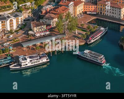 Vista aerea del porto con barche passeggeri ormeggiate e in movimento sull'acqua turchese, circondata da edifici storici e alberi lussureggianti a Riva del G Foto Stock