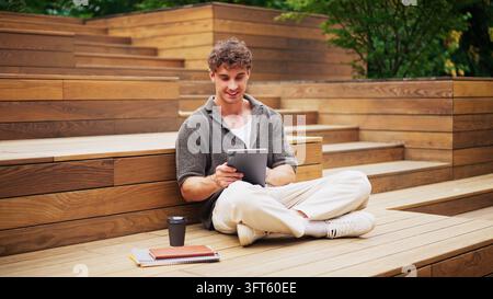 Giovane uomo seduto su gradini di legno all'aperto mentre utilizza un tablet digitale per compiti di lavoro remoto Foto Stock