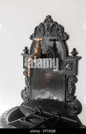 Una vista ravvicinata angolata di una fontana da giardino a parete in ghisa verniciata nera in stile vintage che mostra un rubinetto dell'acqua in ottone antico e decorazioni a rilievo ornate Foto Stock