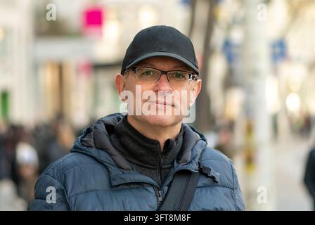 Un uomo di mezza età con un cappello nero e occhiali si trova su una strada cittadina sfocata. Foto Stock
