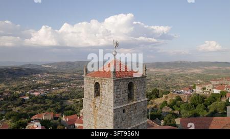 Vista aerea della torre dell'orologio in pietra del villaggio di Monsanto, con tetto a tegole rosse e weathervane a forma di gallo, che si affaccia sul paesaggio rurale di Mo Foto Stock