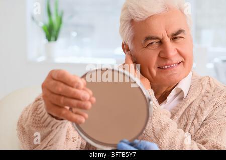 Perdita uditiva e salute degli anziani, uomo anziano che indossa un apparecchio acustico guardandosi allo specchio per una migliore comunicazione e qualità della vita a casa Foto Stock