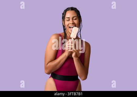 Gioia estiva e stile di vita da vacanza, giovane donna felice che mangia un ghiacciolo in costume da bagno su sfondo viola per il marketing stagionale e il tempo libero Foto Stock