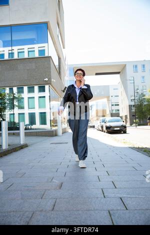 Una donna d'affari matura sorridente parla al telefono mentre tiene un laptop in una strada della città di Helsinki fuori da un ufficio moderno Foto Stock