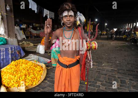 Sadhu con una fascia arancione e collane di perle si trova su ciottoli di pietra di notte vicino a un mucchio di fiori gialli, Bengala Occidentale, Kolkata, India Foto Stock