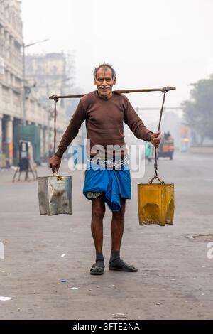 Un uomo sorridente in maglione marrone e lungi blu tiene un'asta di bambù con un contenitore di metallo ad ciascuna estremità, Bengala Occidentale, Kolkata, India Foto Stock