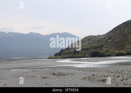 vallata fluviale intrecciata, letto ghiaioso ampio, più canali poco profondi, contrafforti boscosi scuri, picchi lontani (grandangolo, panorama, cielo nuvoloso, movimento Foto Stock