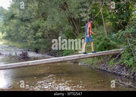 Vista laterale di un escursionista maschio che cammina lungo un stretto ponticello di legno sopra il fiume Maniavka nel villaggio di Maniava, Carpazi, Ucraina. Equilibrio e avventura Foto Stock